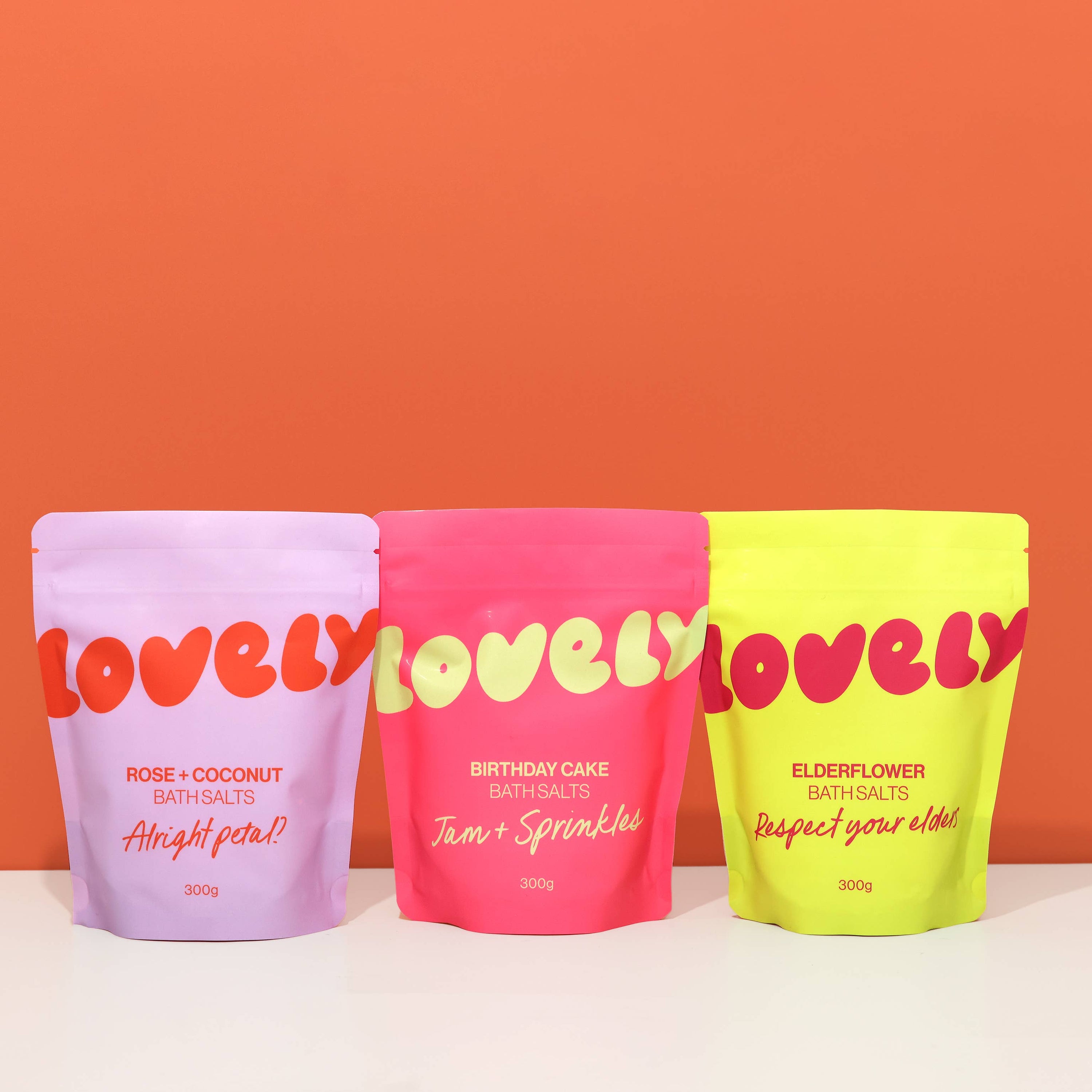 Three colorful 'lovely' bath salts packages against an orange background