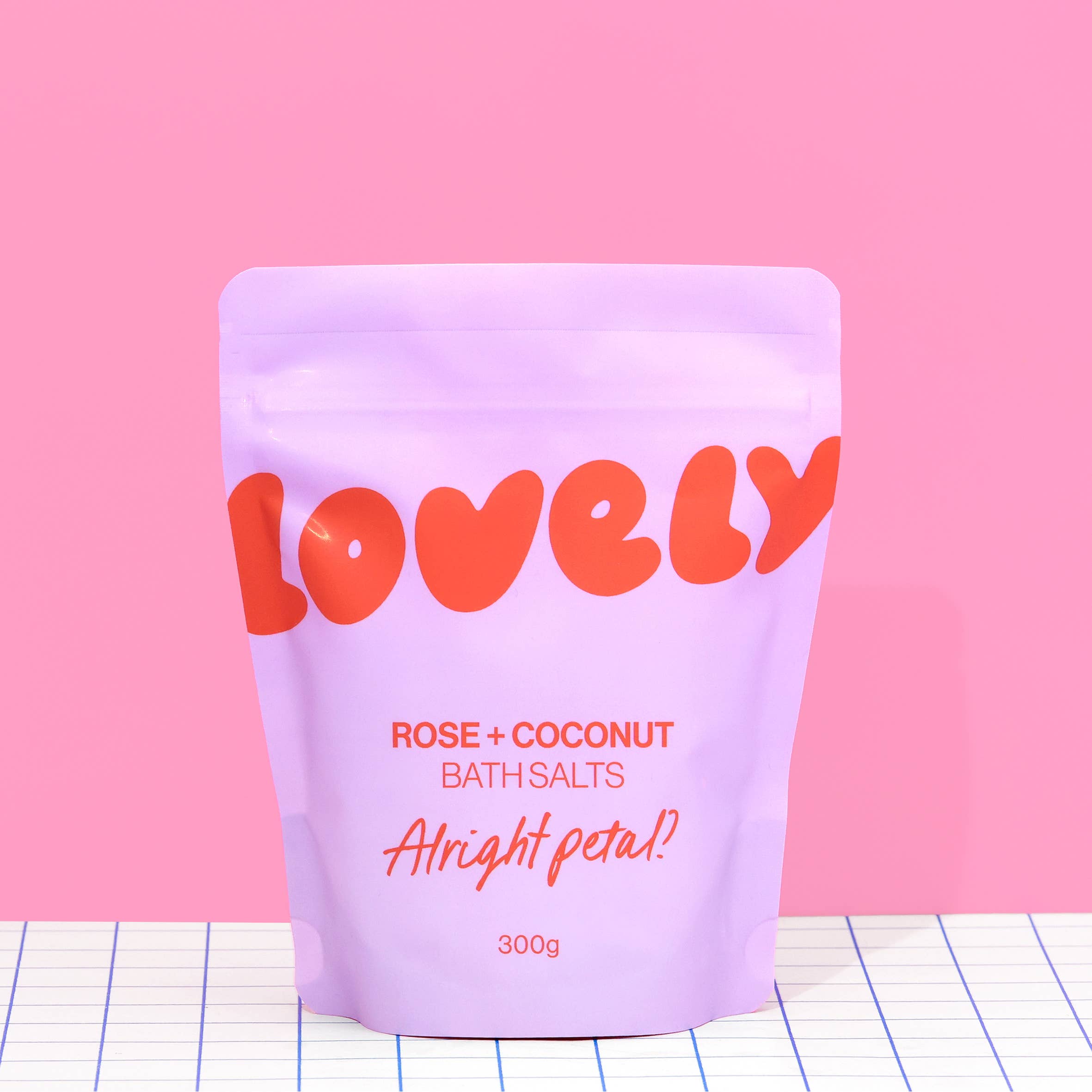 Purple packaging of 'Lovely' rose and coconut bath salts on a pink background