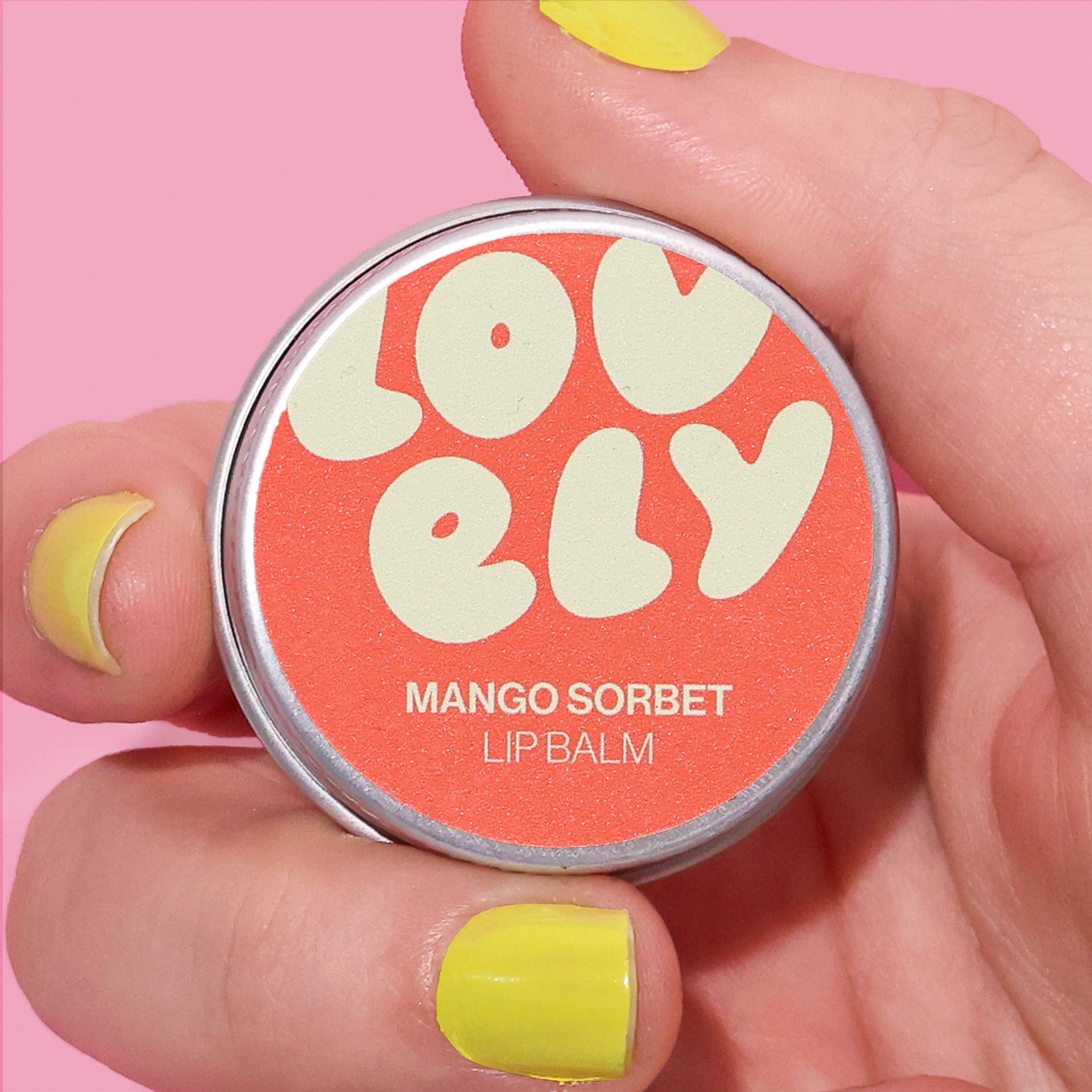 Lip balm container labeled 'Lovely Mango Sorbet' held by a hand with yellow nail polish on a pink background.