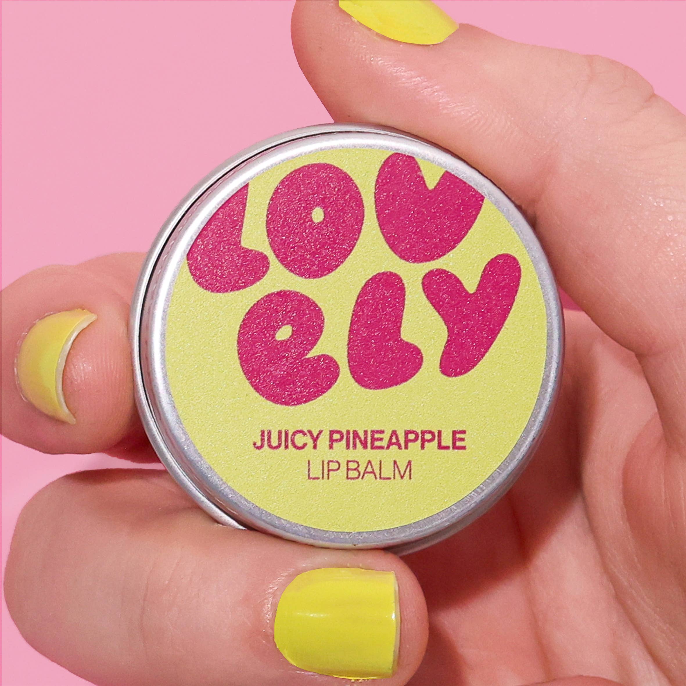 Lip balm container labeled 'Lovely' with 'Juicy Pineapple' flavor held by a hand with yellow nail polish on a pink background.
