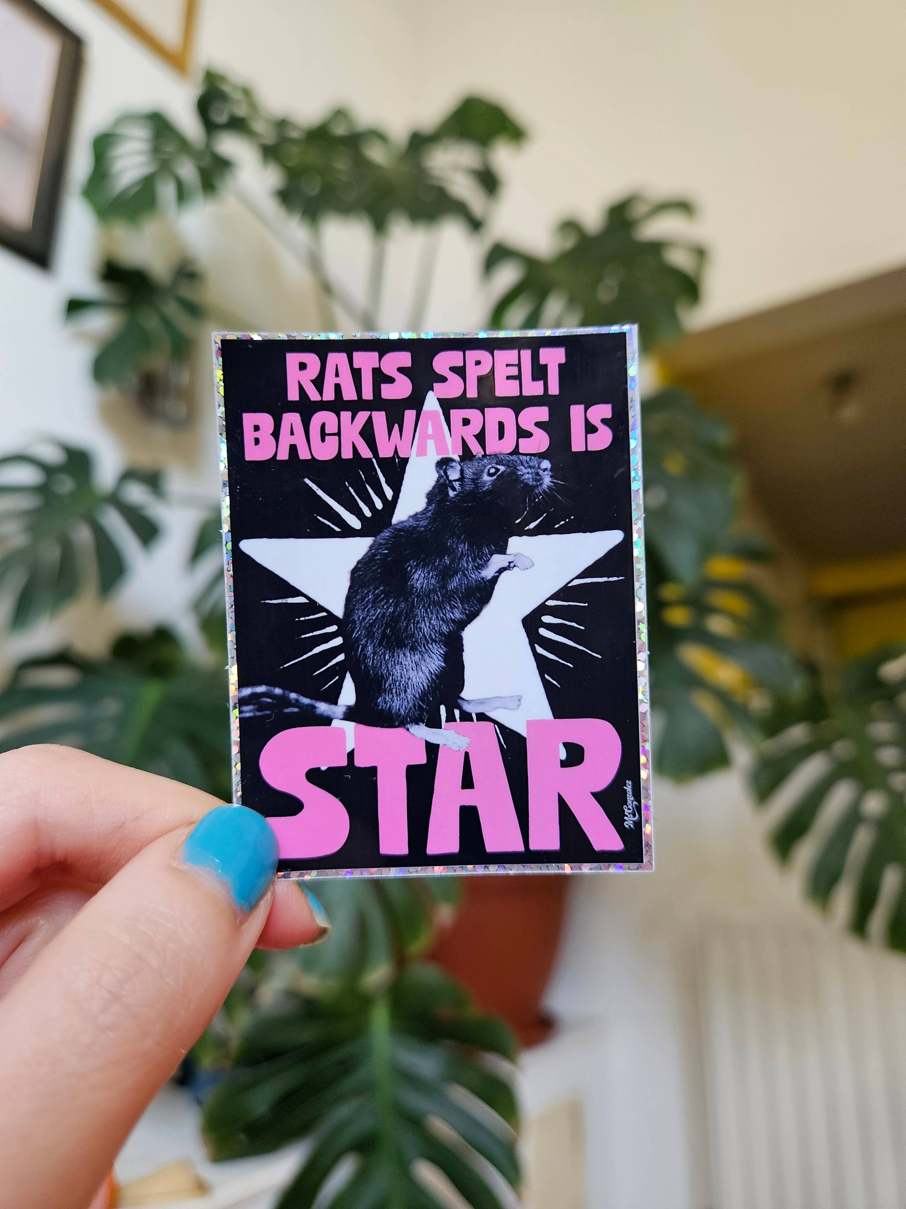 Sticker with text 'RATS SPELT BACKWARDS IS STAR' held by a hand in front of green plants.