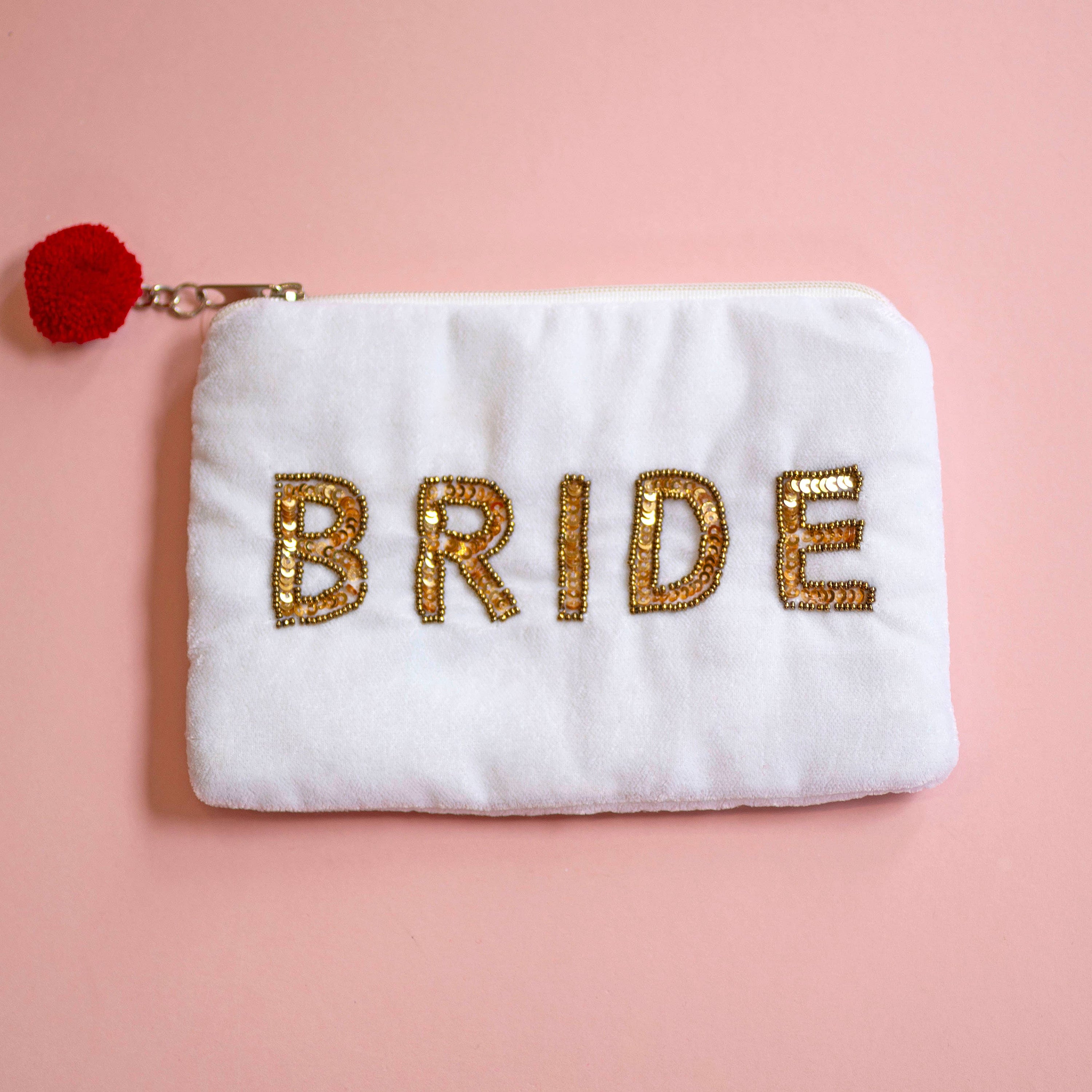 A white plush zipper pouch with the word 'BRIDE' embroidered in gold sequins, featuring a small red pom-pom charm on the zipper pull, set against a solid pink background.