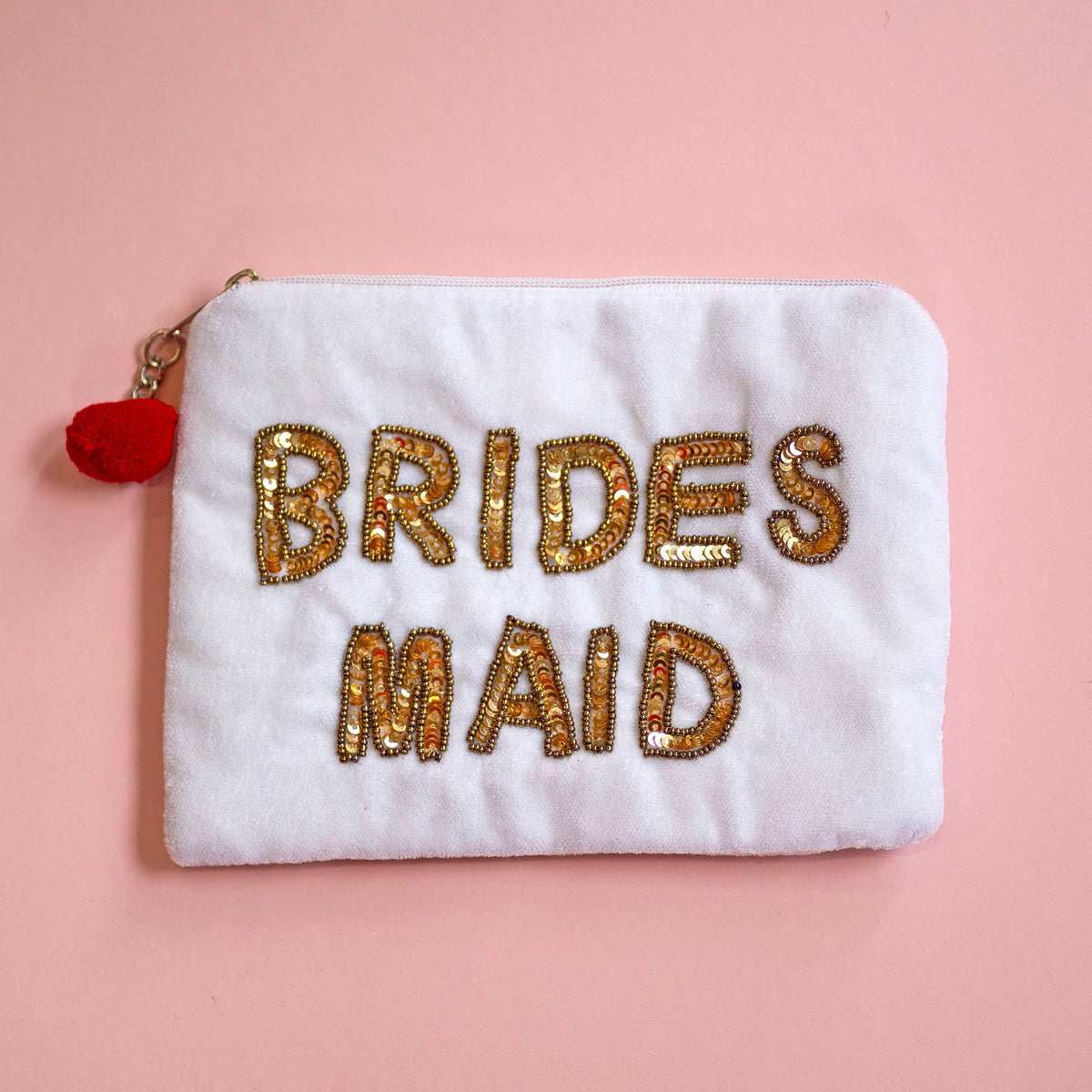 A white plush zipper pouch with 'BRIDES MAID' embroidered in gold sequins, featuring a small red pom-pom charm on the zipper pull, set against a solid pink background.