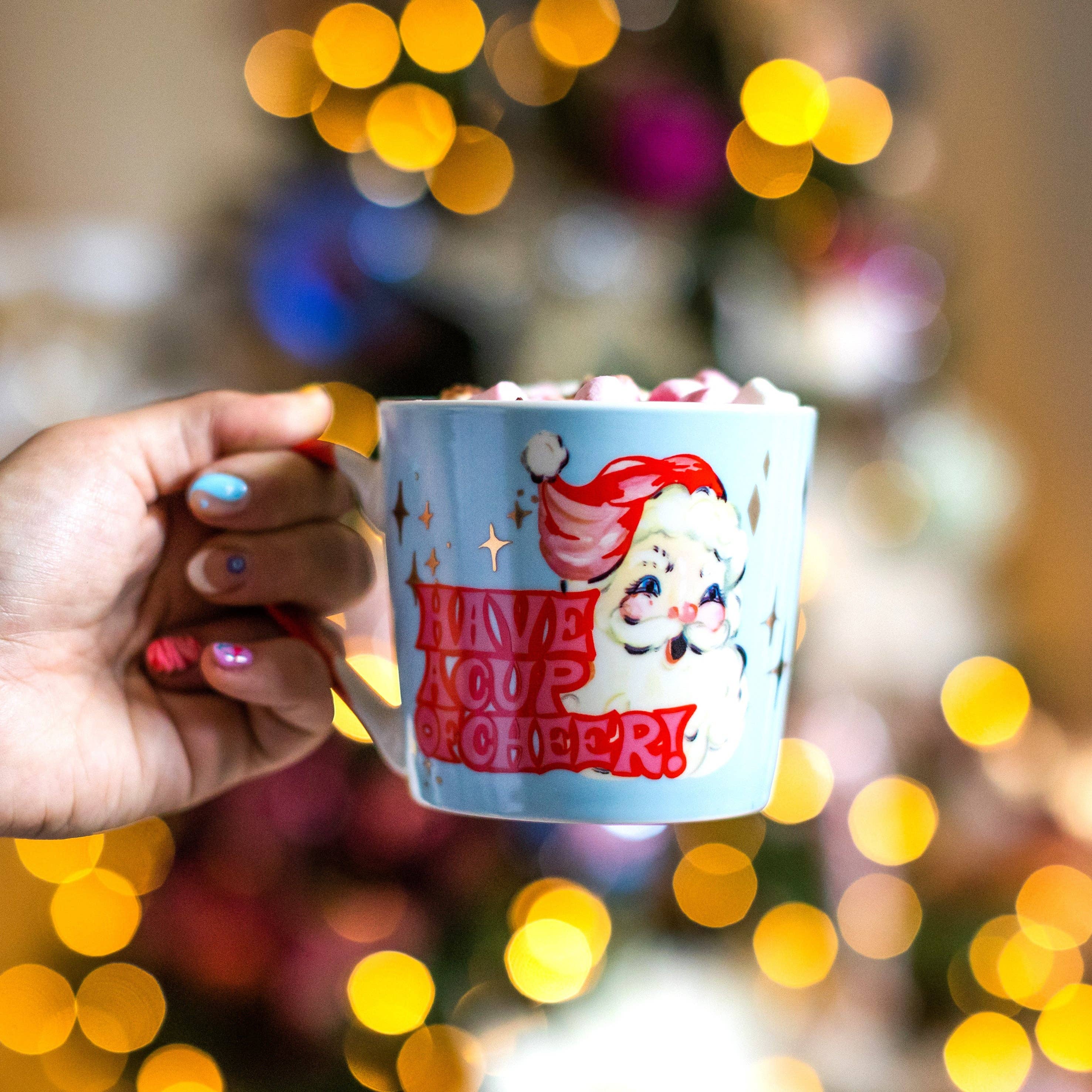 Hand holding a mug with Santa Claus design and 'Have a cup of cheer' text against a festive background.