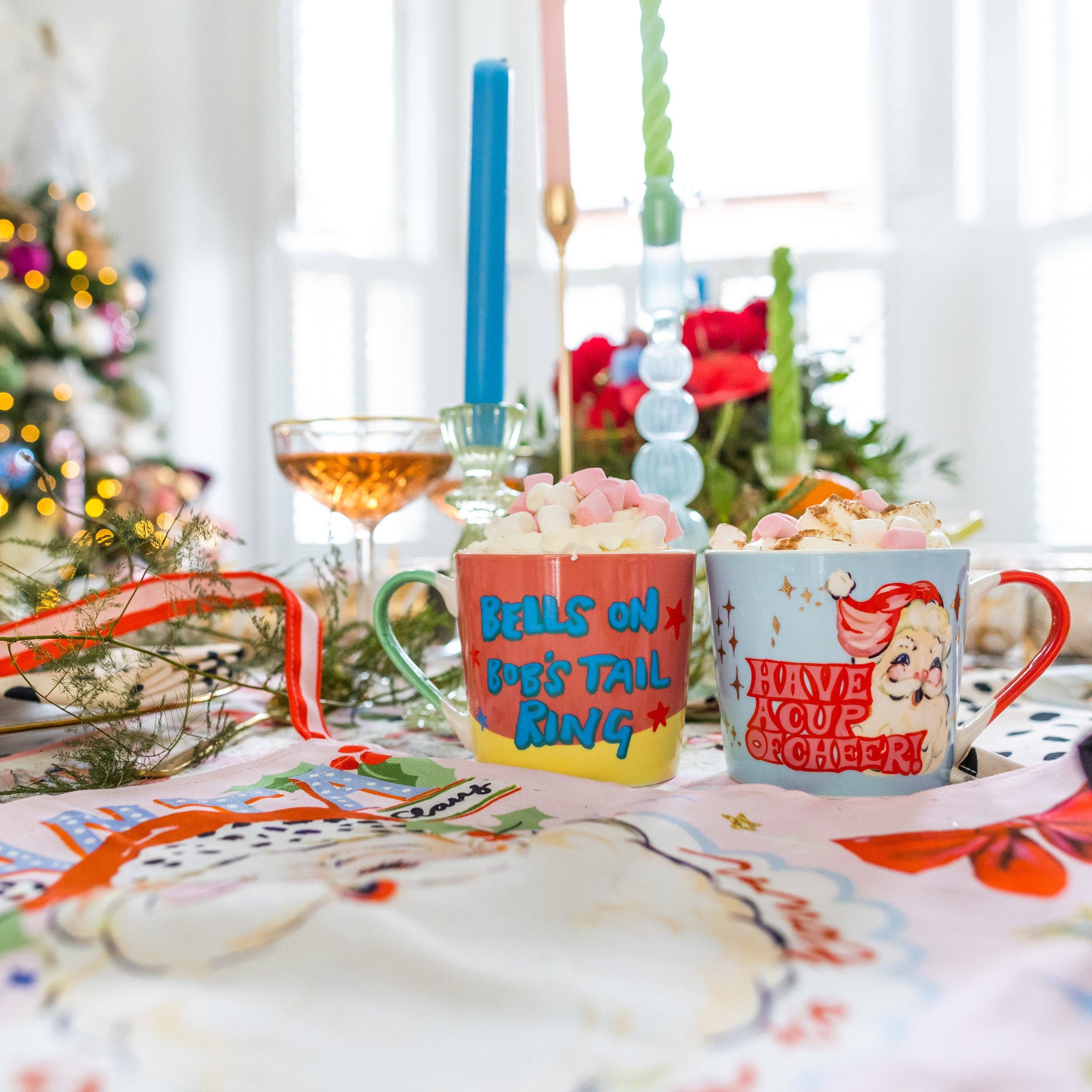 Two Christmas-themed mugs on a festive table with candles and decorations.