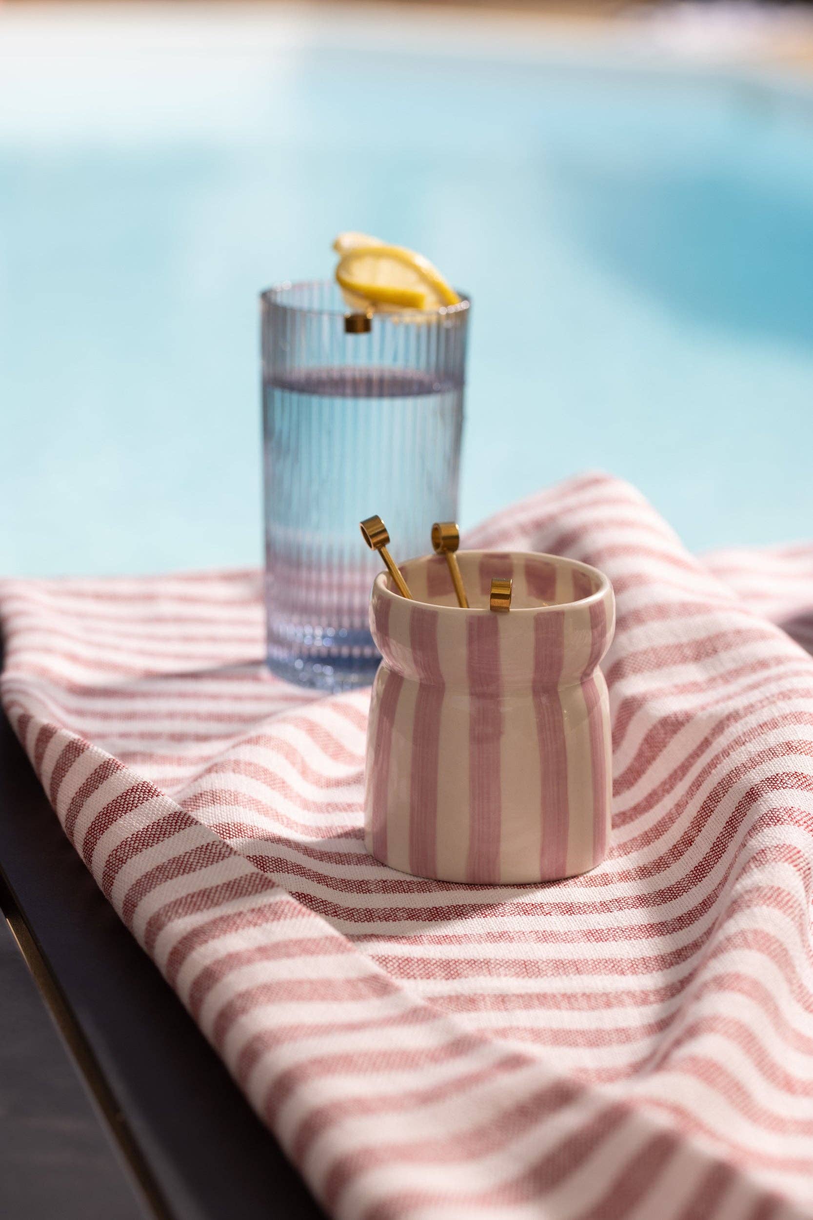 Glass of water with lemon and a striped towel by a pool by the striped ceramic candle displayed on it, being used as a lass after it was a candle. 