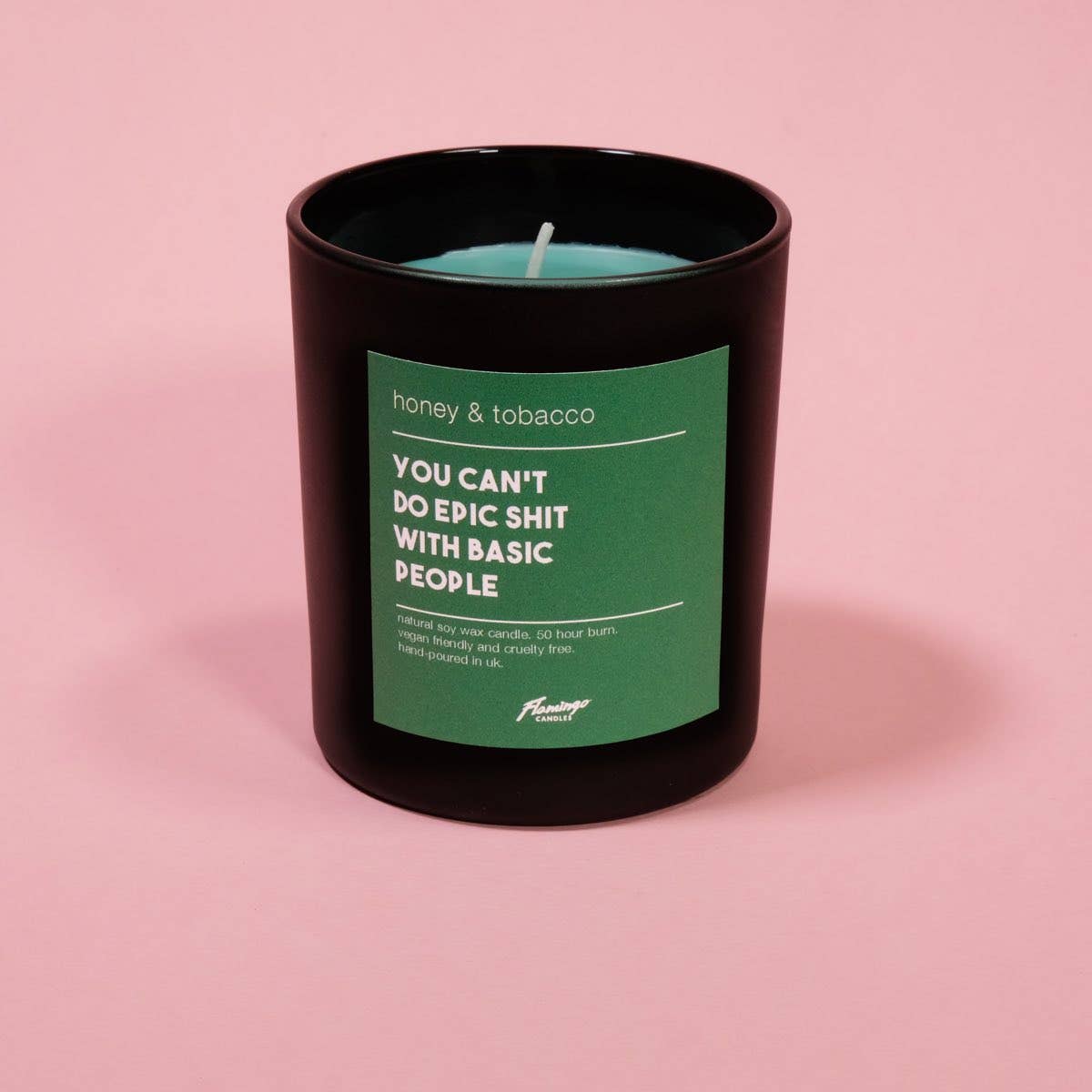 Scented candle in a black jar container with a green label on a pink background. Candle sticker reads "You can't do epic shit with basic people"