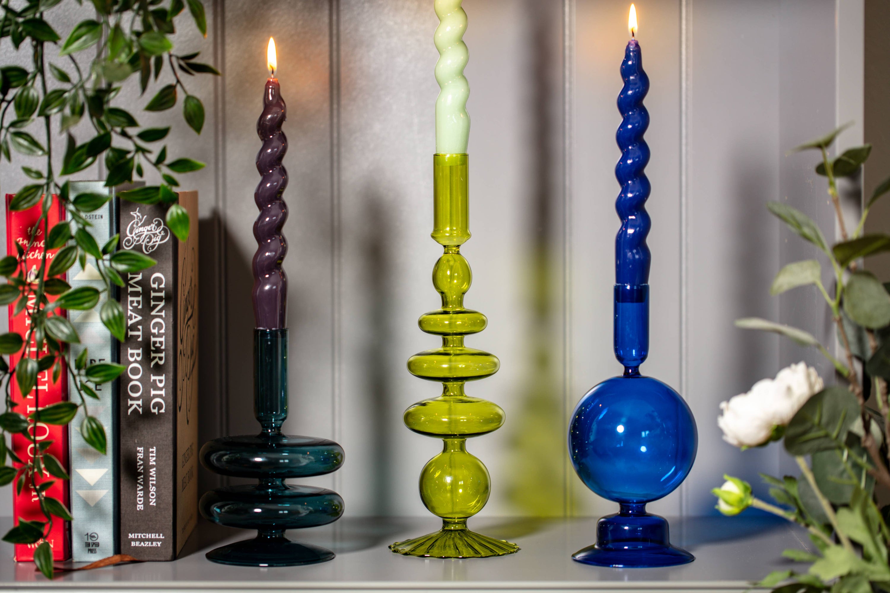 Colorful twisted candlesticks on a shelf with books and plants.