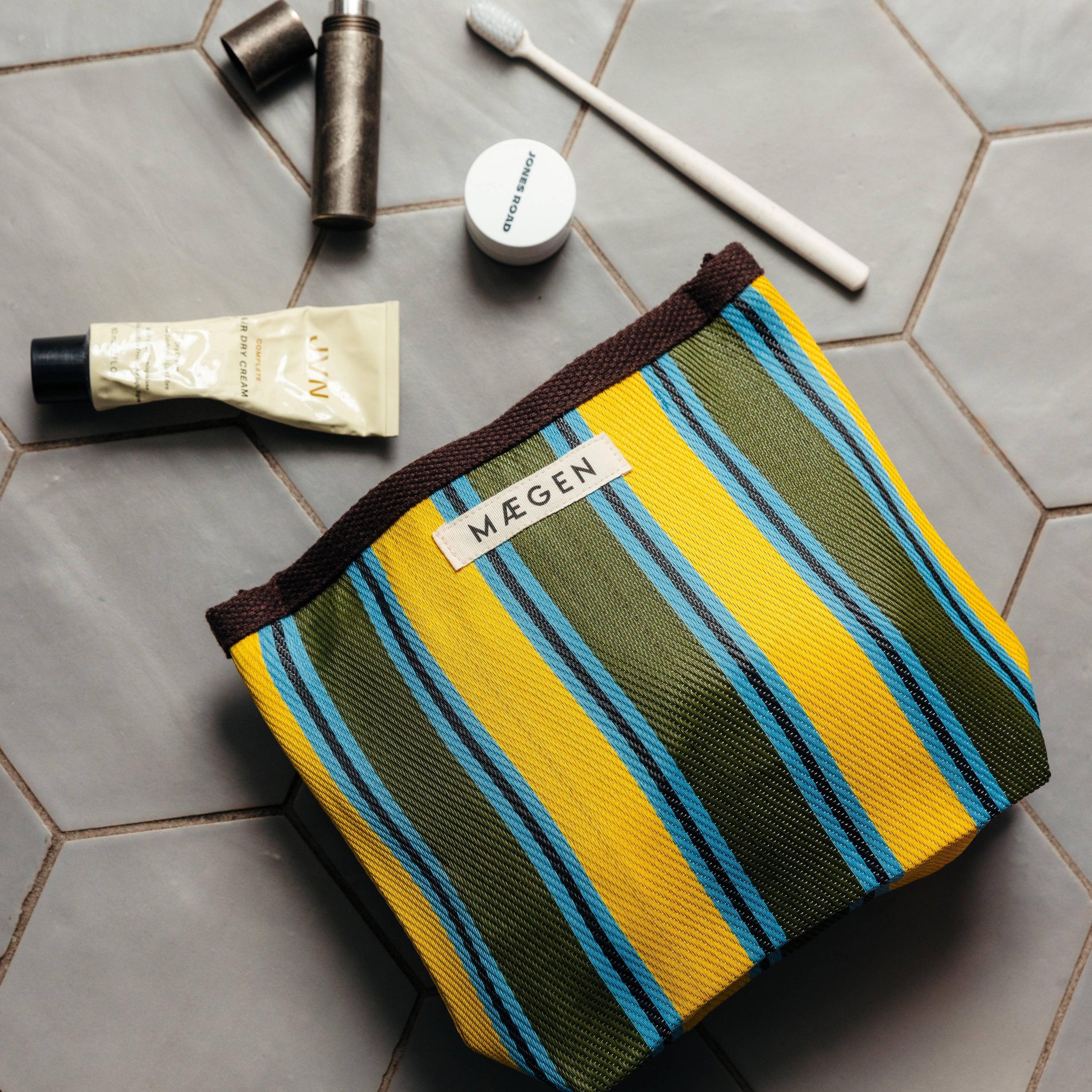 Striped toiletry bag with 'MAEGEN' label on a tiled surface with bathroom items.