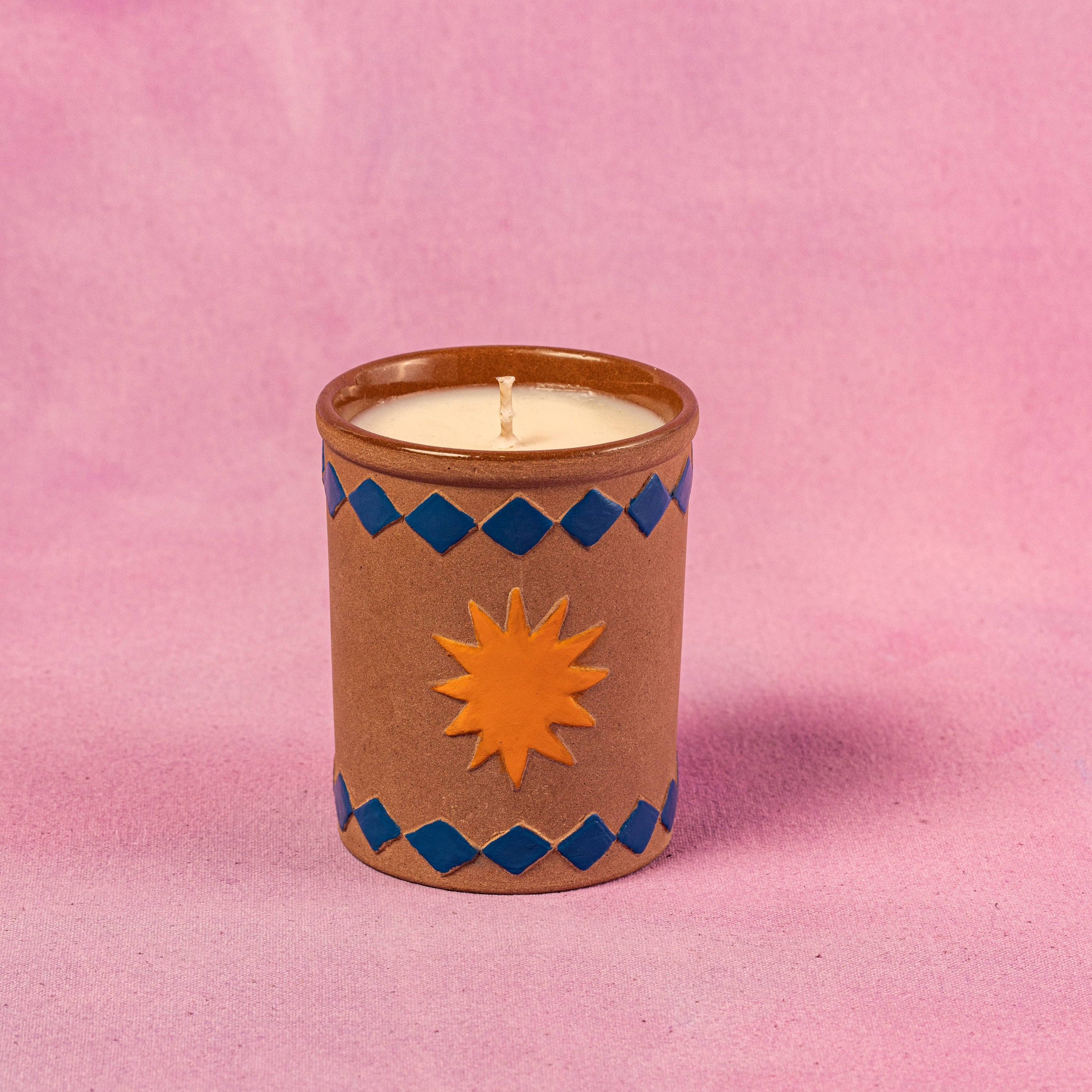 Candle in a decorative brown container with blue patterns and an orange sun design on a pink background