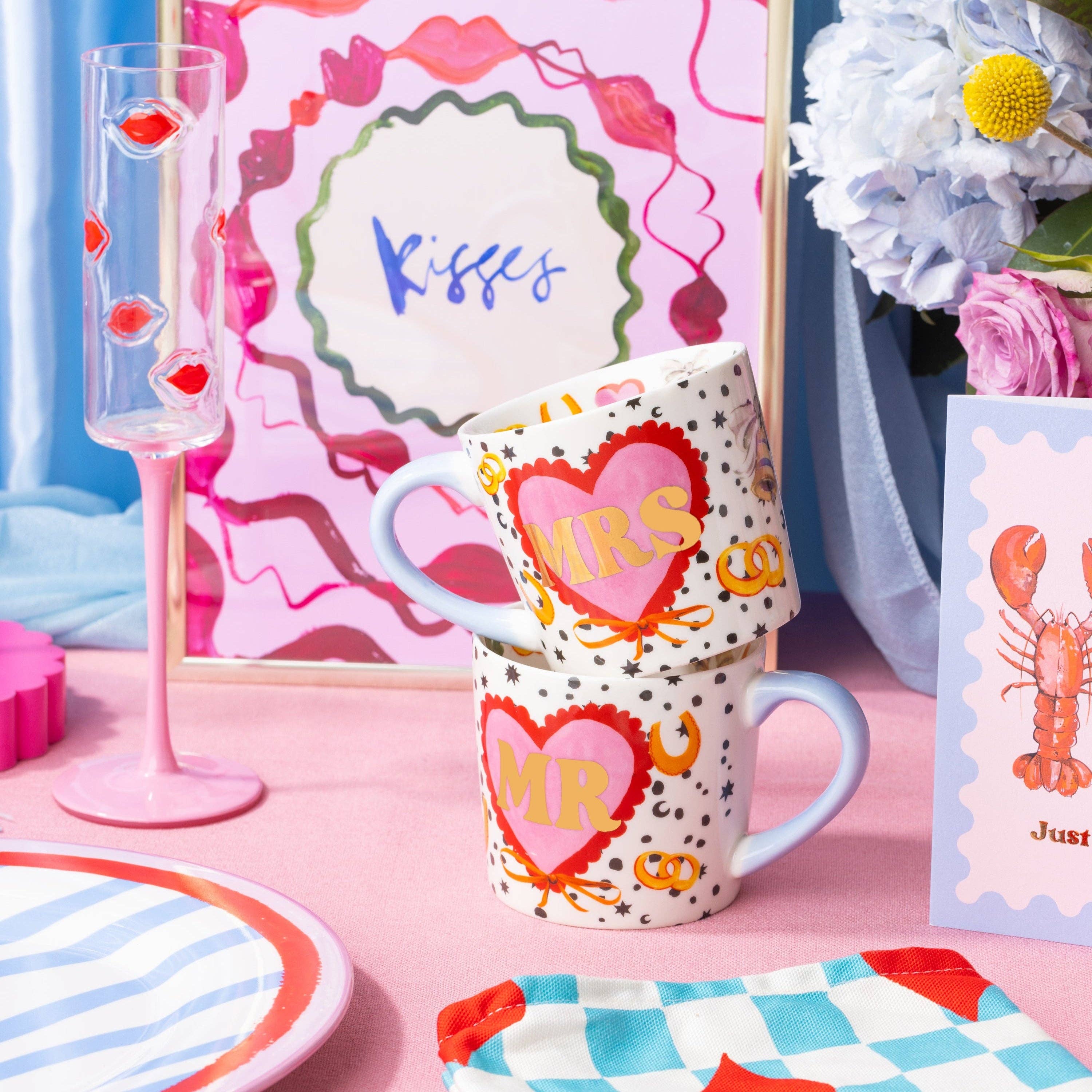 Decorative table setting with mugs, glasses, and cards on a pink surface.