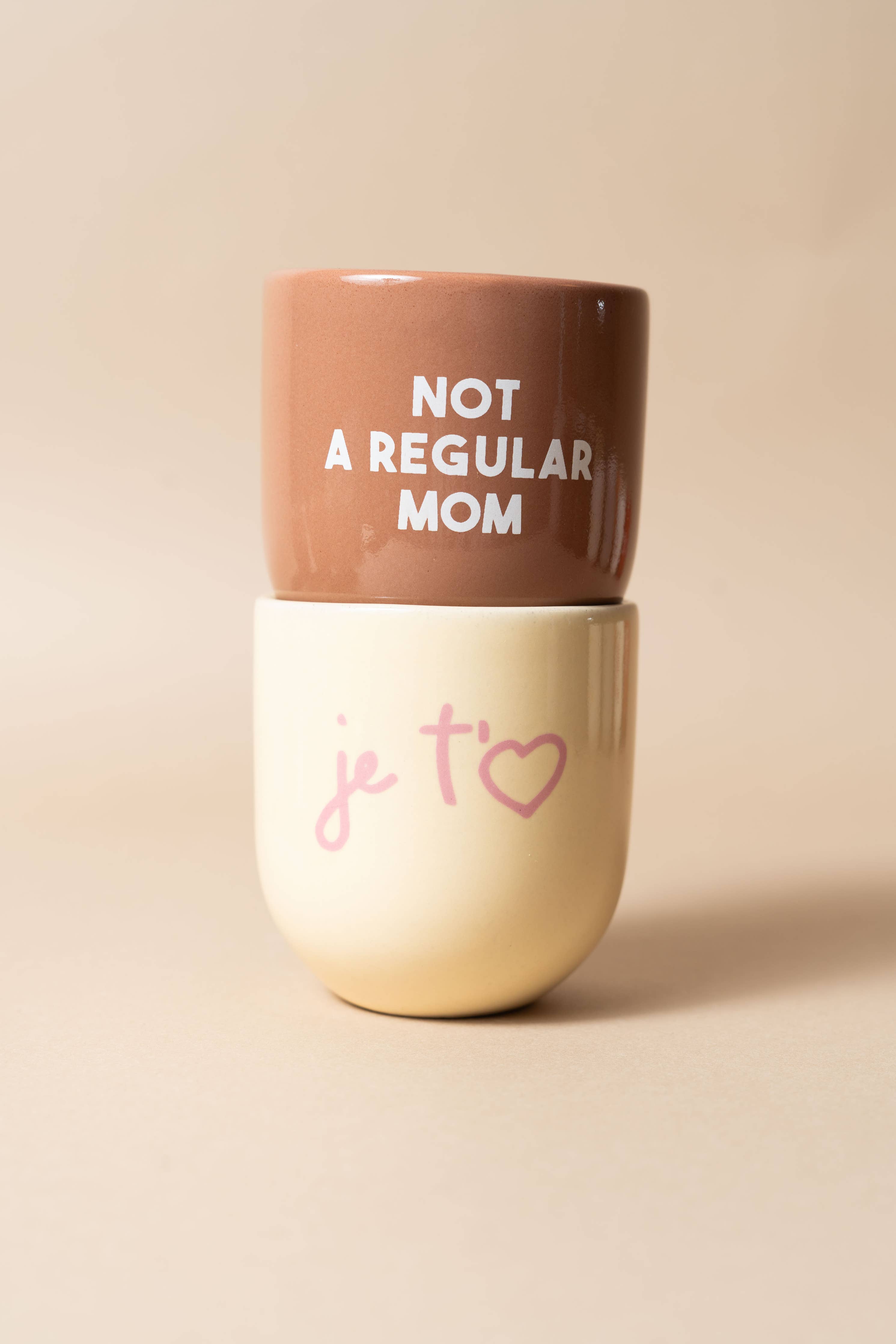 Two tumblers stacked on a beige background, with one saying 'Not a Regular Mom' and the other 'je t§é'