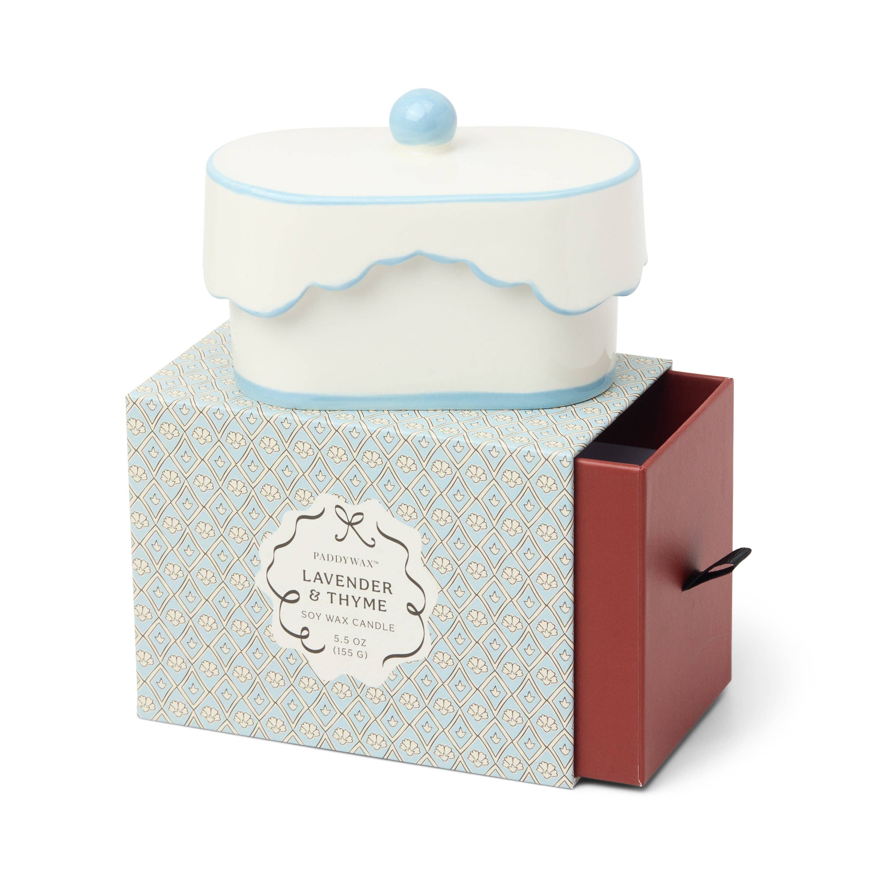 A scented candle in a decorative cake-shaped container with a blue lid on its special packaging