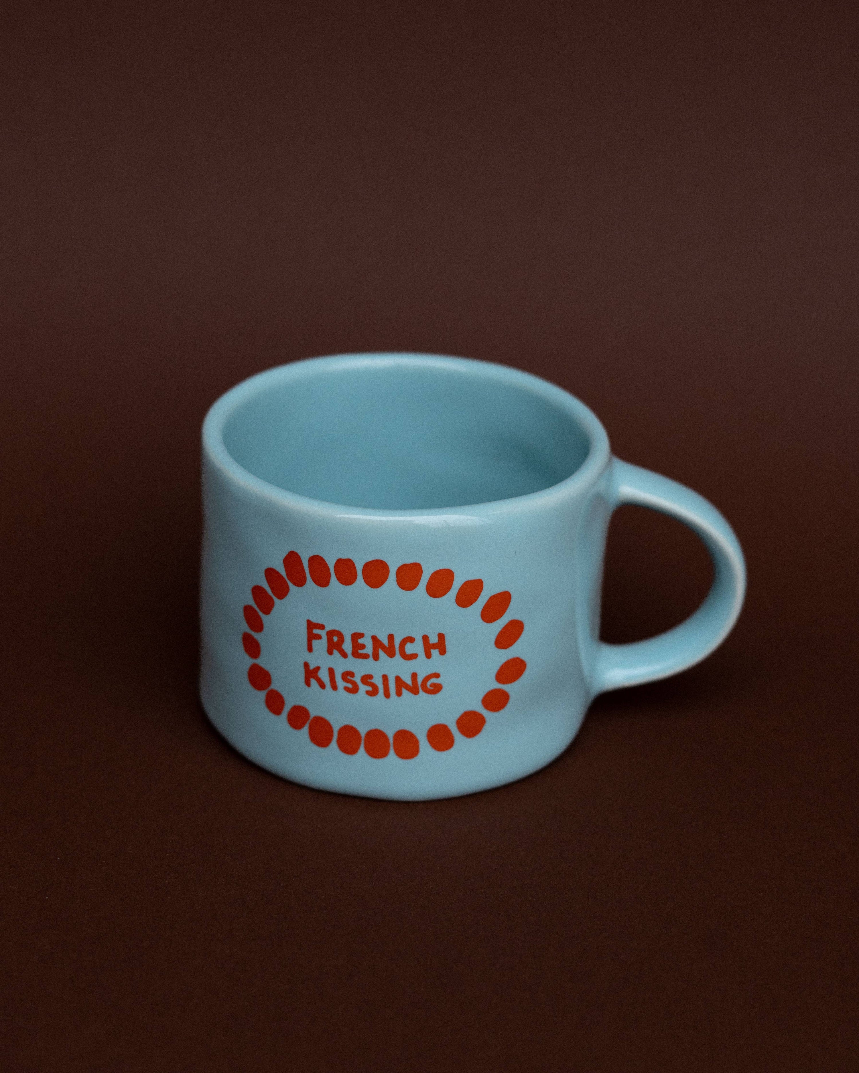 Light blue mug with red text 'French Kissing' on a brown background