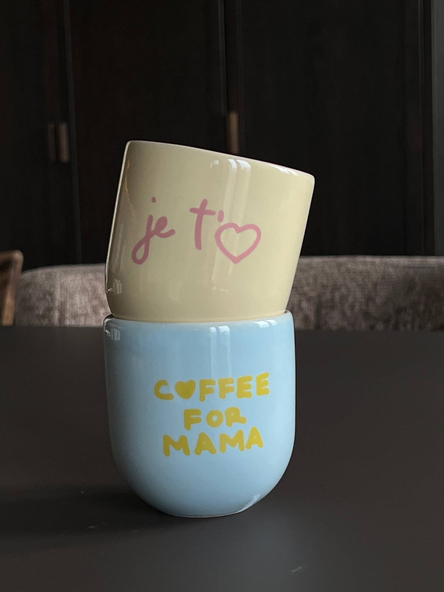 Two ceramic cups stacked on a dark surface, one beige with 'je tàime' and one blue with 'COFFEE FOR MAMA'.