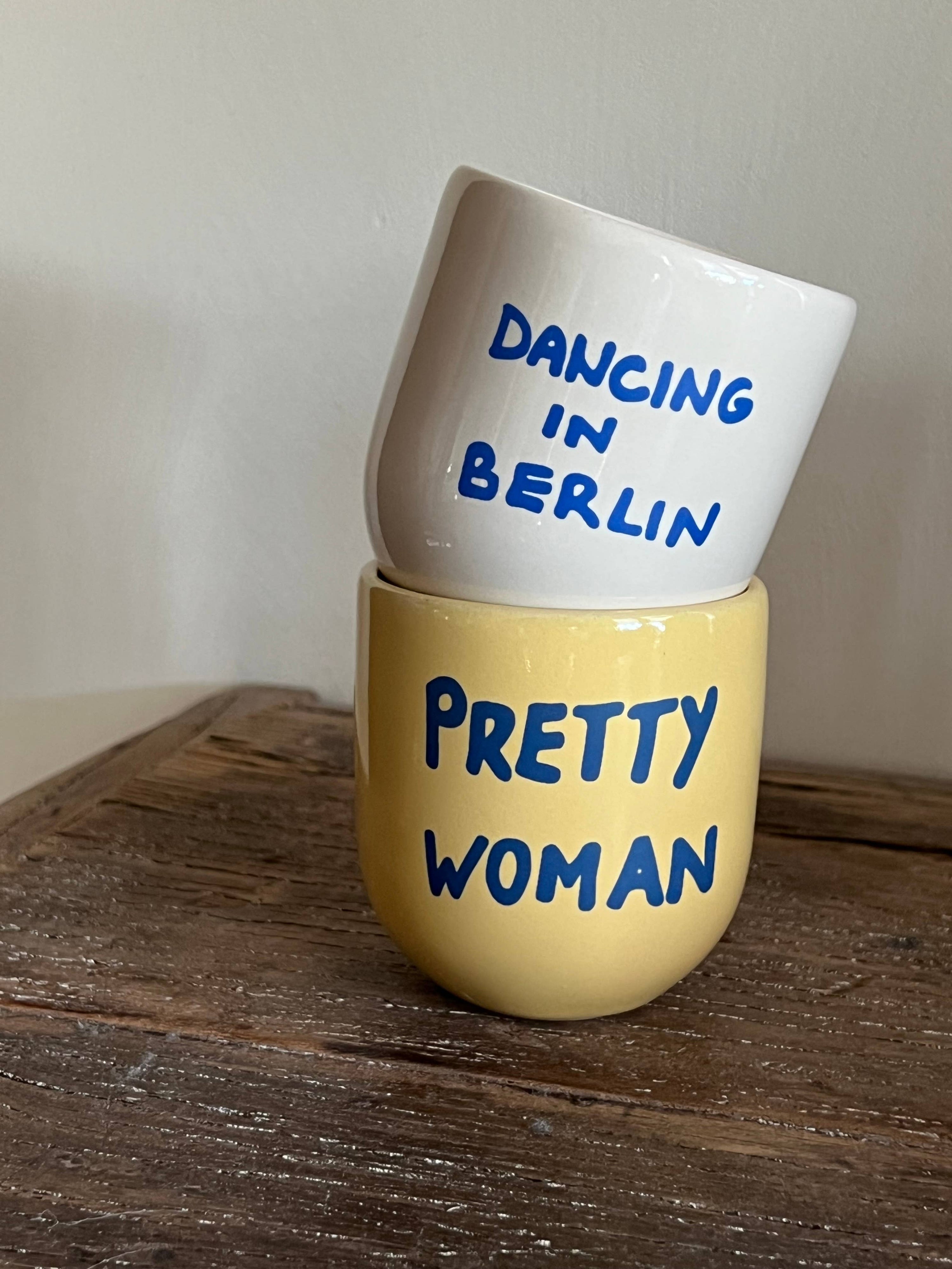 Two ceramic cups stacked on a wooden surface with text 'Dancing in Berlin' and 'Pretty Woman'.