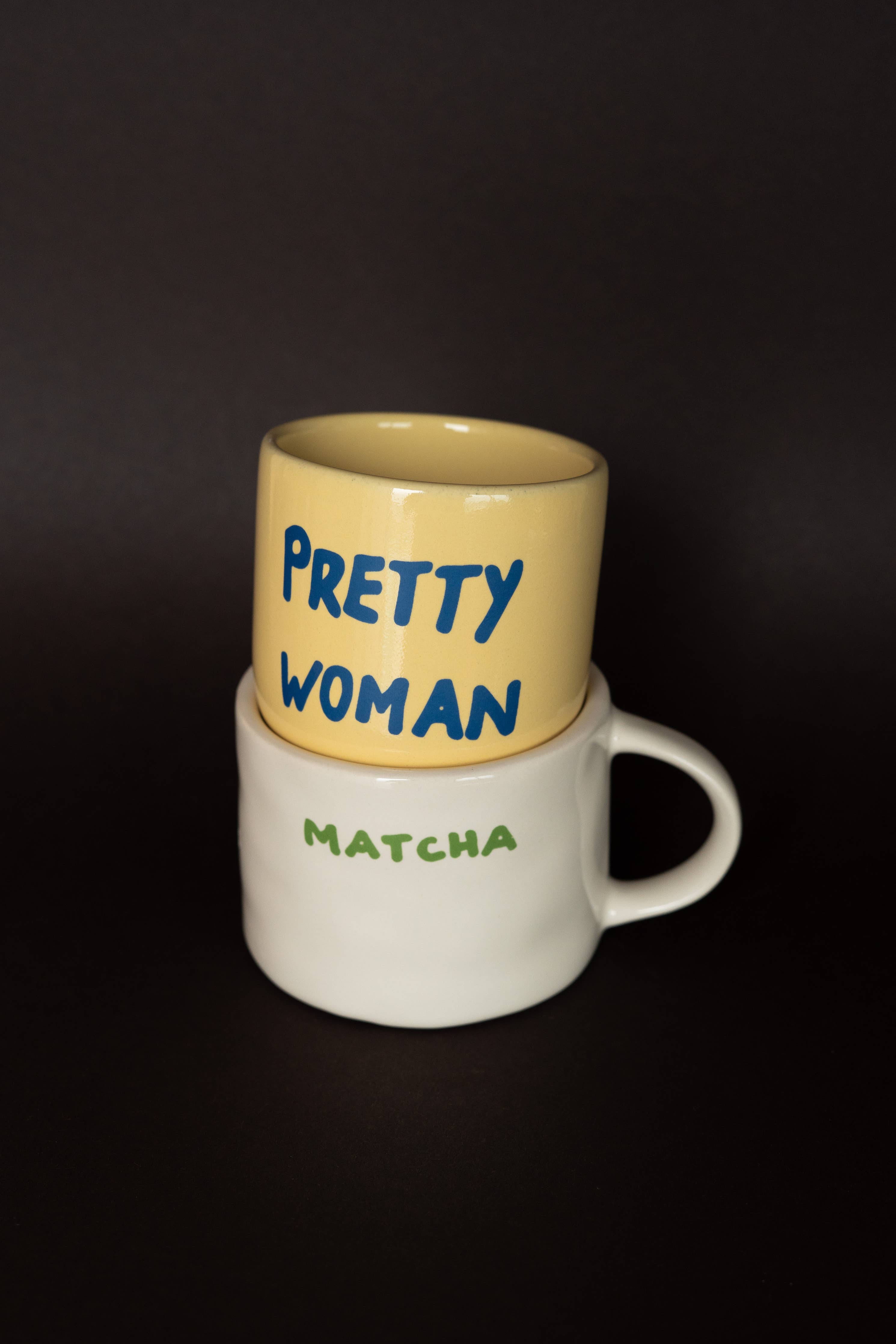 Two mugs stacked on a dark background, one yellow with 'Pretty Woman' text and one white with 'Matcha' text.