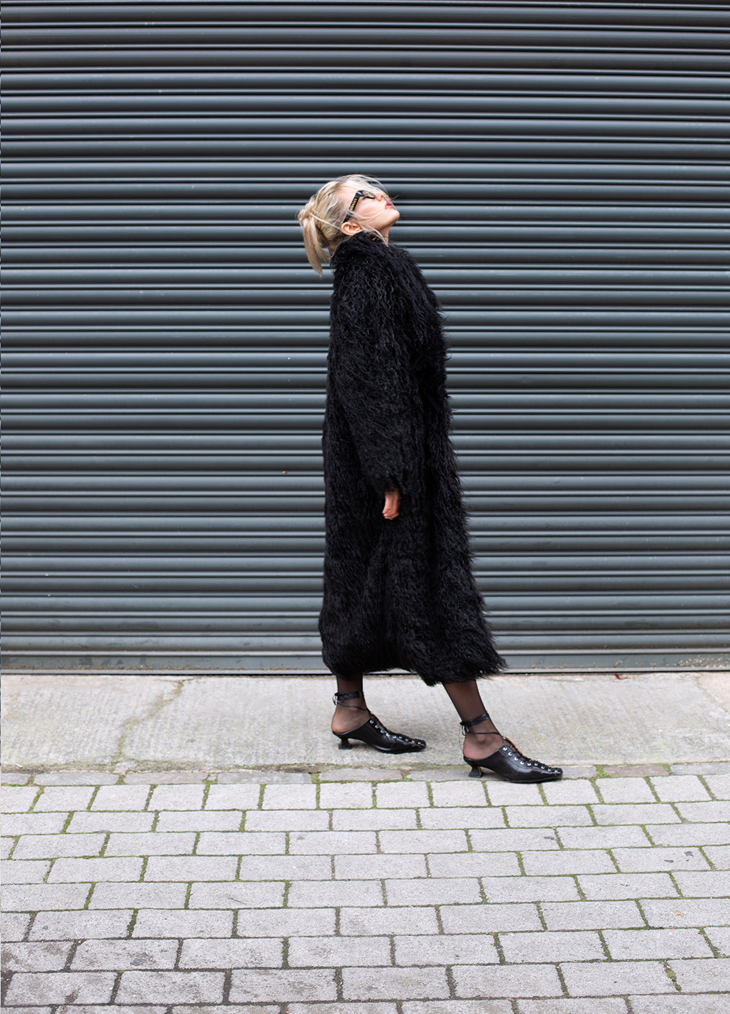 Person wearing a long black coat standing in front of a gray shutter.