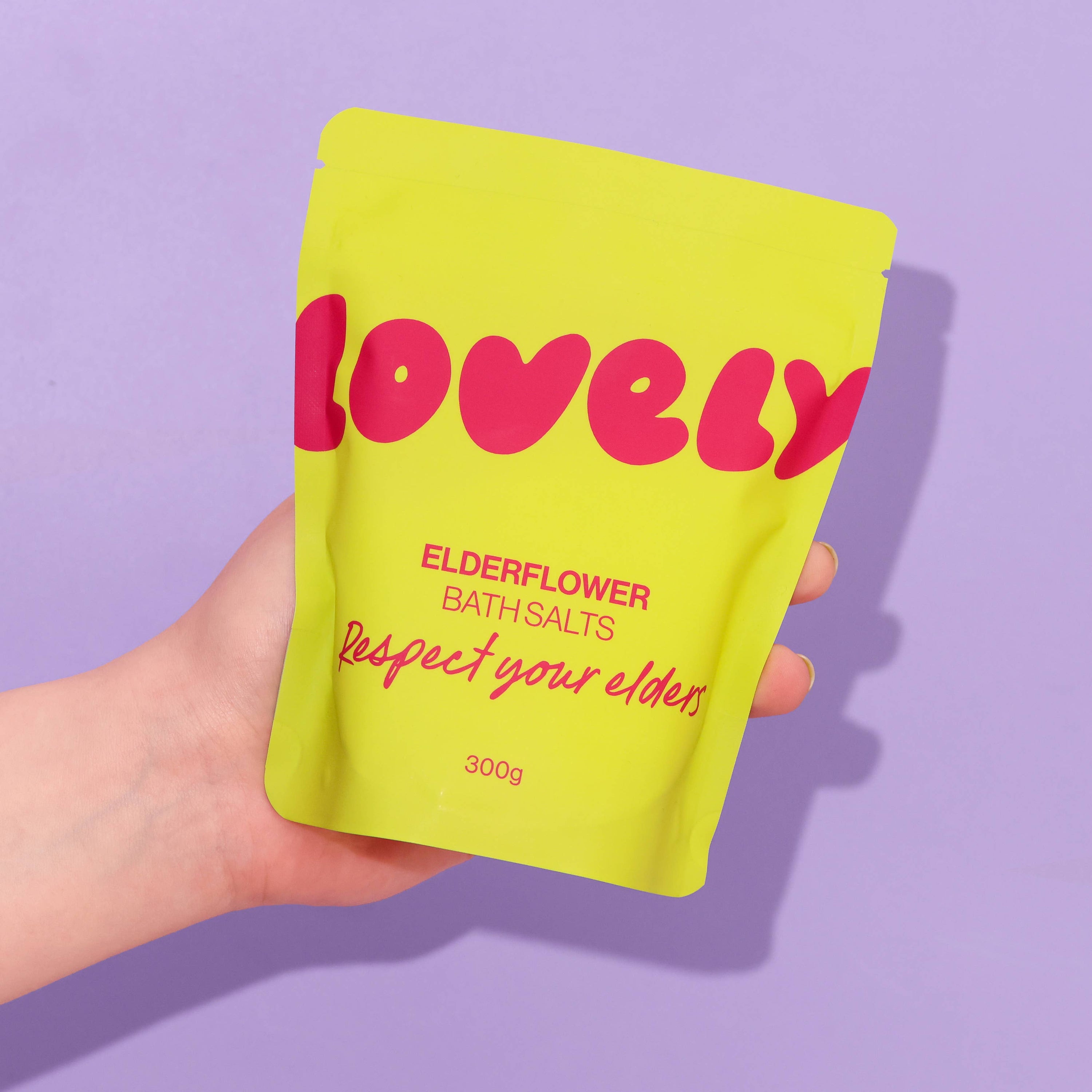 Hand holding a yellow package of 'Lovely' elderflower bath salts on a purple background