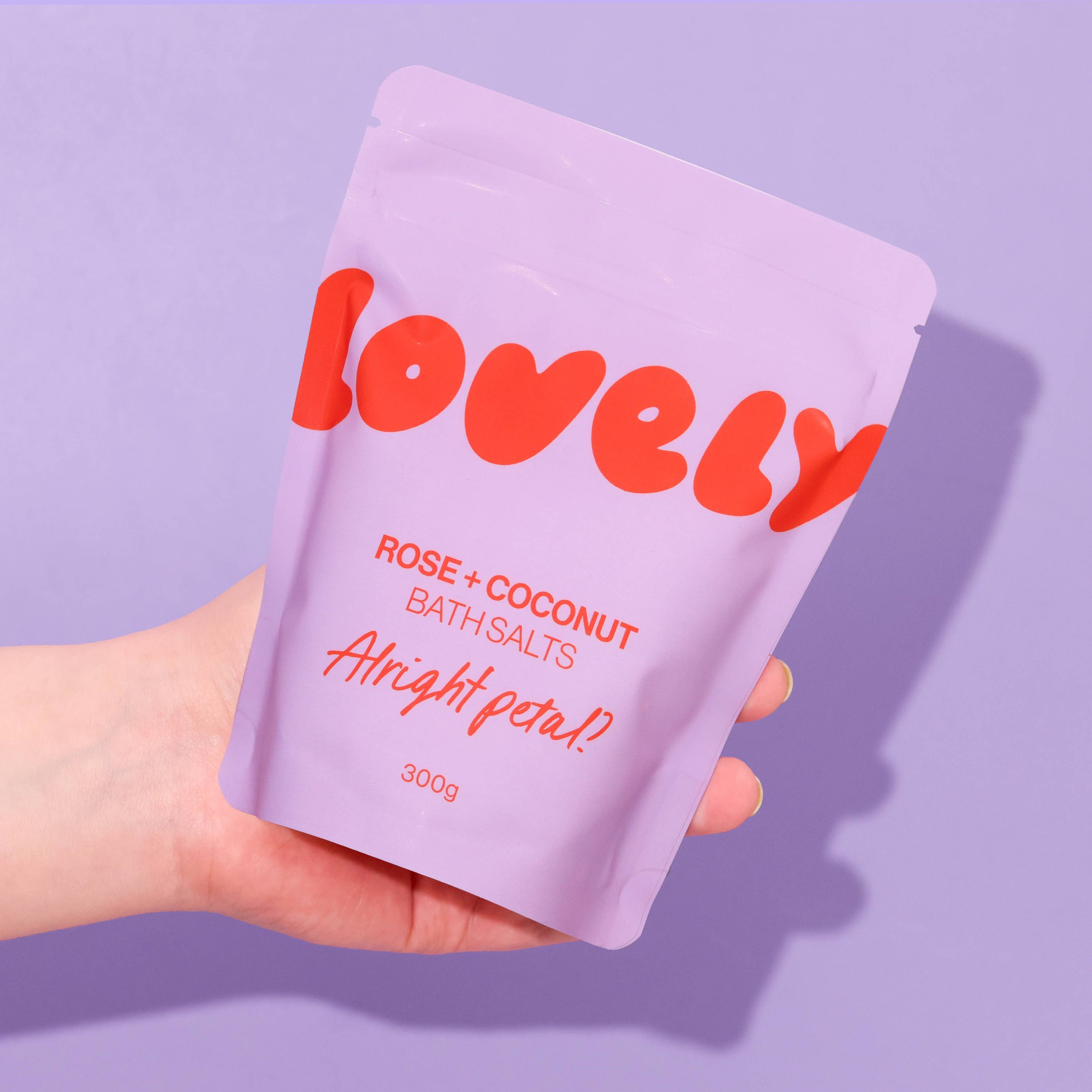Hand holding a purple packet of 'Lovely' rose and coconut bath salts against a purple background