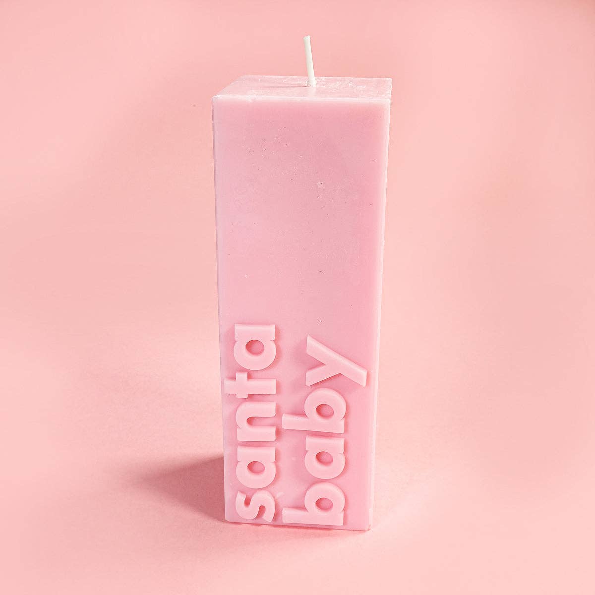 PINK RECTANGULAR PILLAR SLOGAN CANDLE WITH SLOGAN"SANTA B ABY" AGAINST A PINK BACKDROP