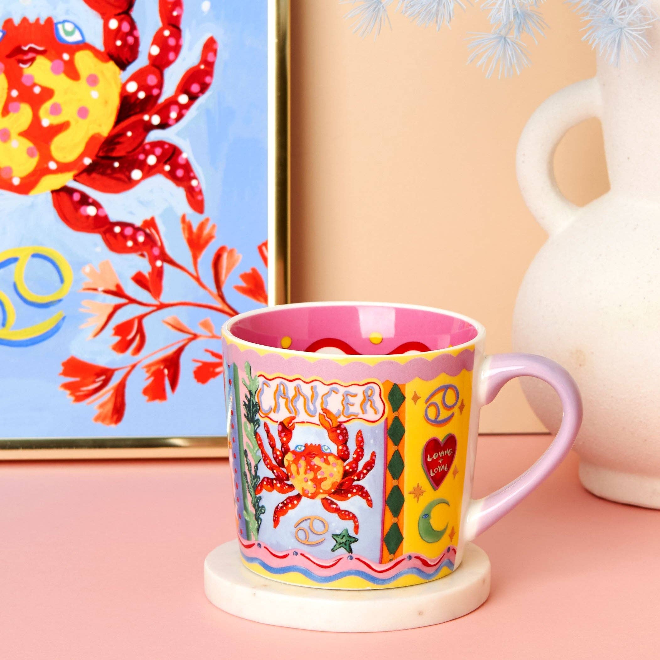 Colourful mug with crab design and 'Cancer' text on a pink surface, with a decorative painting in the background.