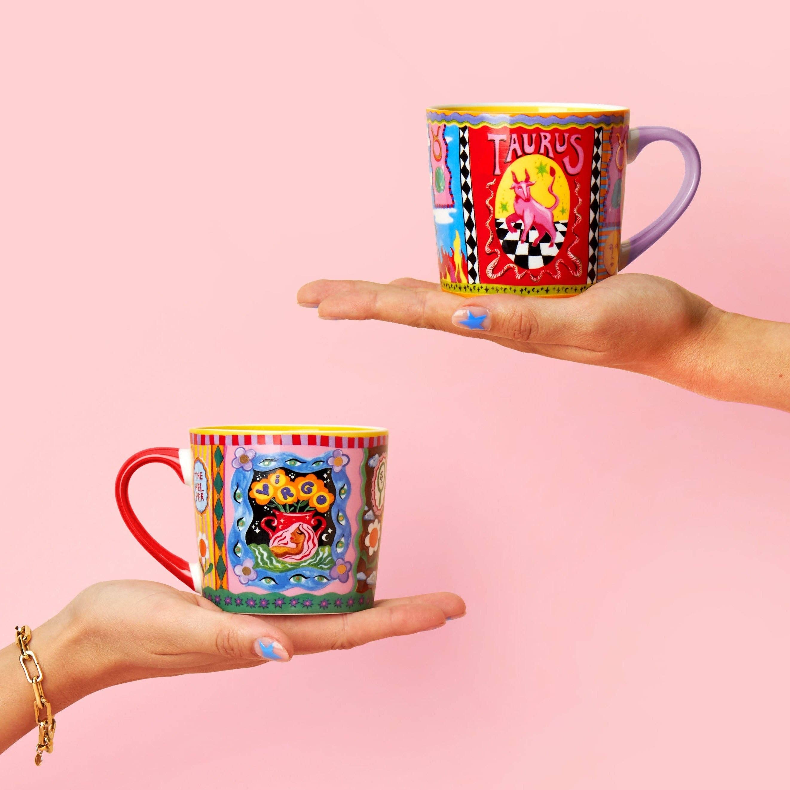 Two colourful mugs with zodiac designs held against a pink background