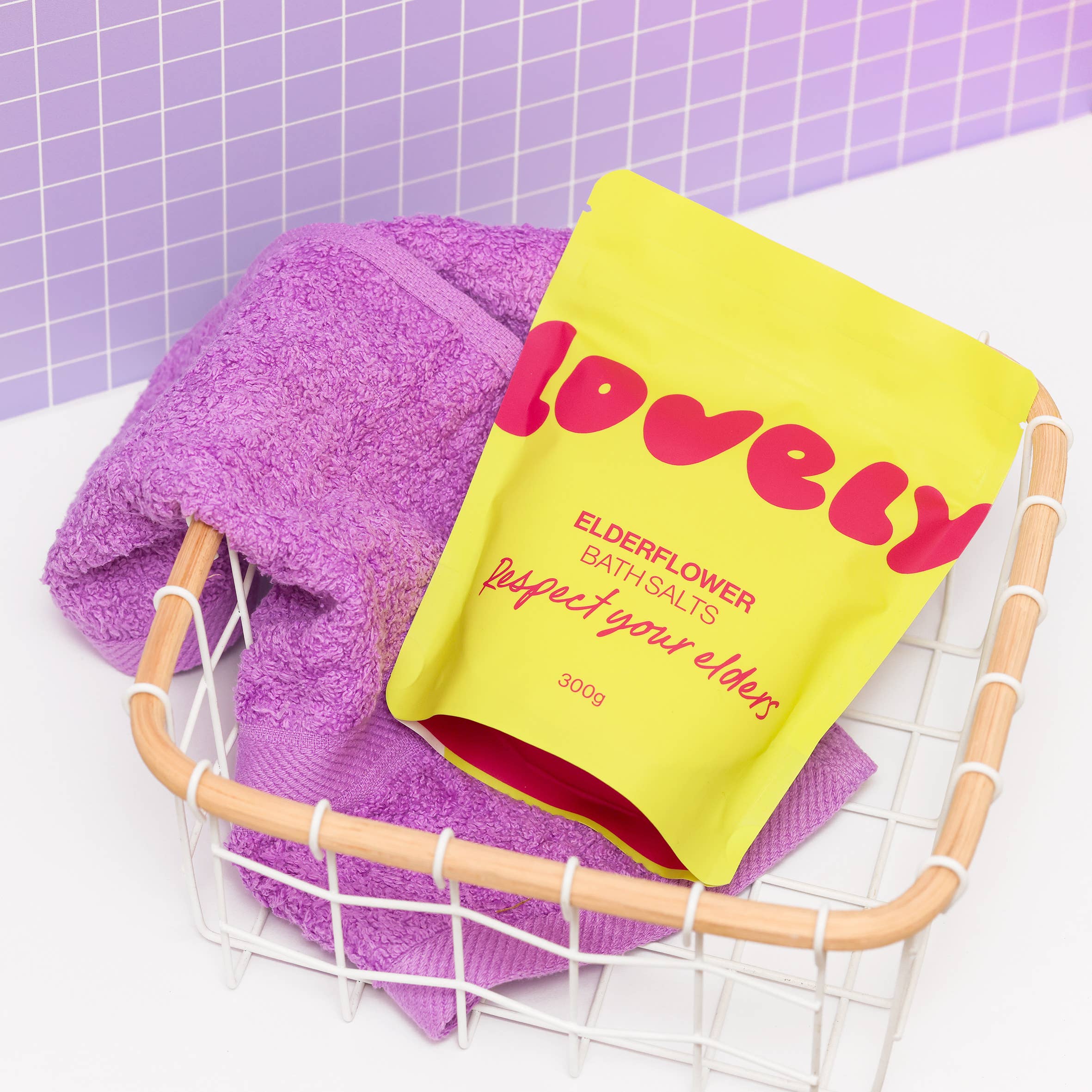 Yellow packet of 'lovely' elderflower bath salts with a purple towel in a basket against a purple tiled wall.
