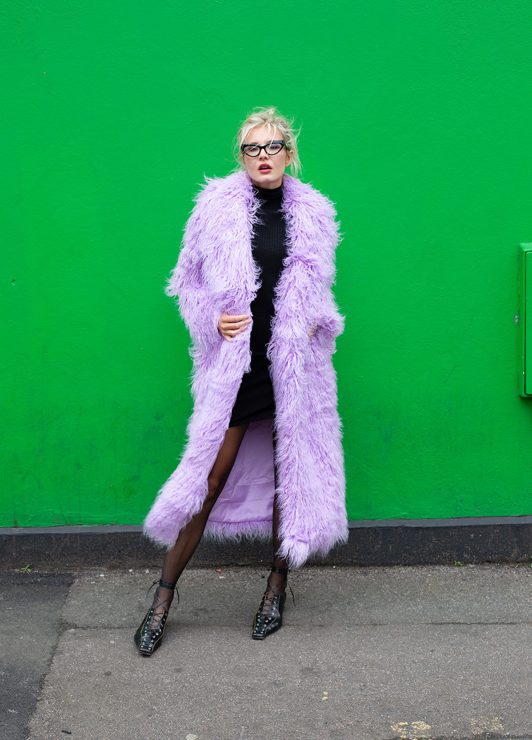 Person wearing a long purple fur coat against a green wall.