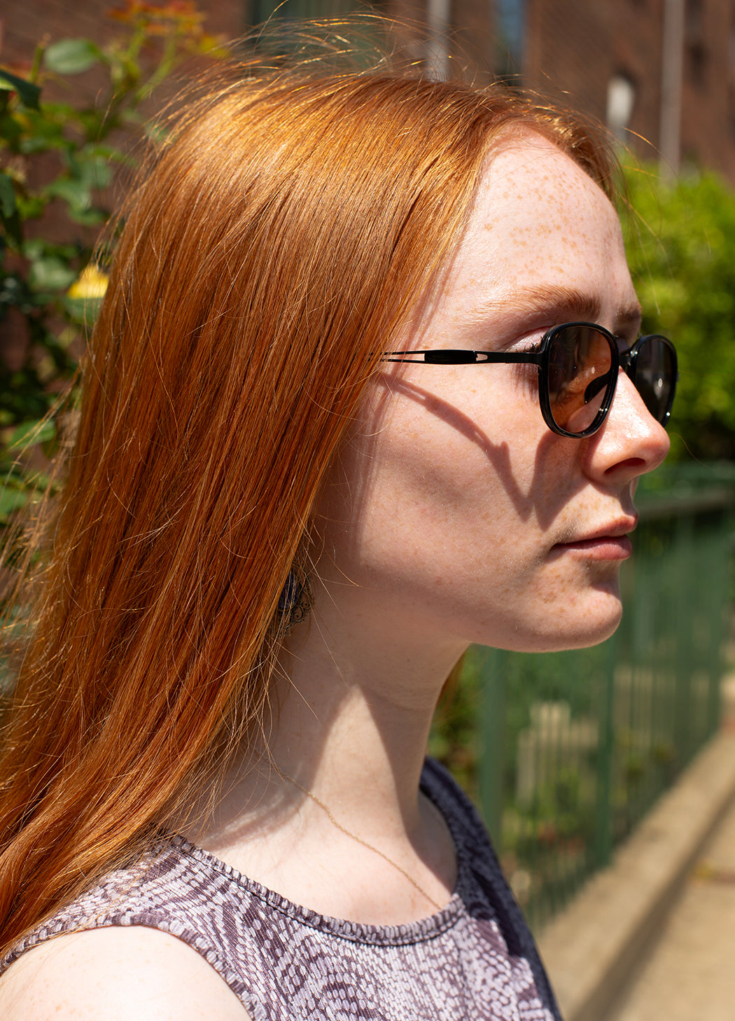 A close-up side profile of a woman with long red hair wearing black sunglasses with dark lenses in an outdoor setting.