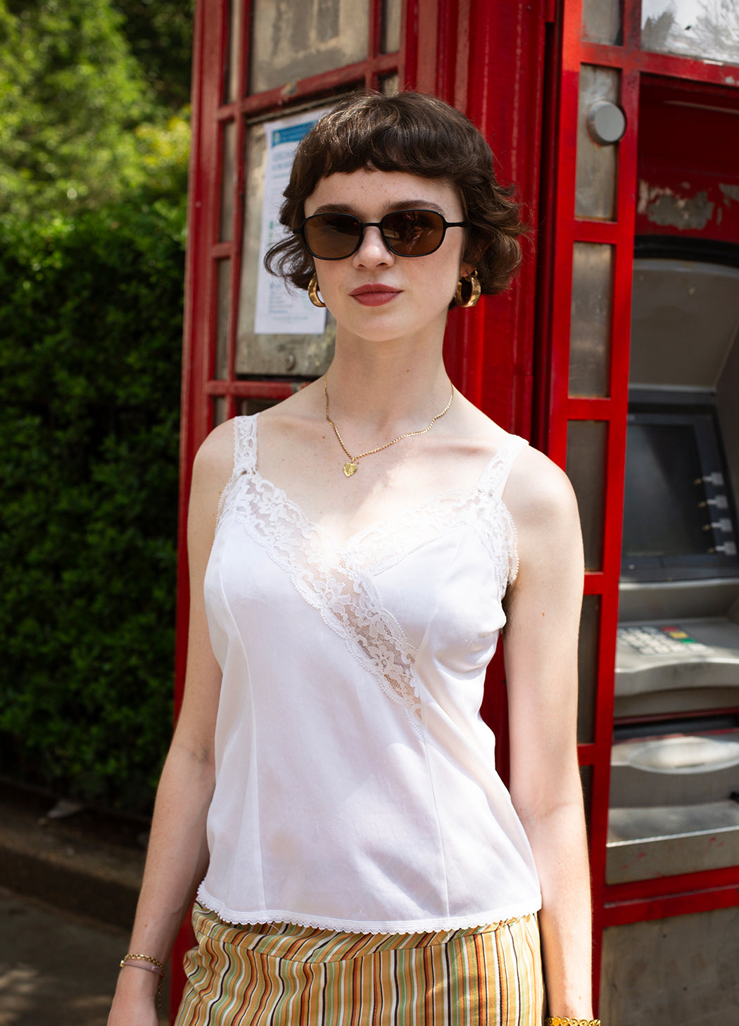 A woman with short dark hair and sunglasses wearing a white lace-trim camisole top and striped trousers, standing in front of a red telephone booth.