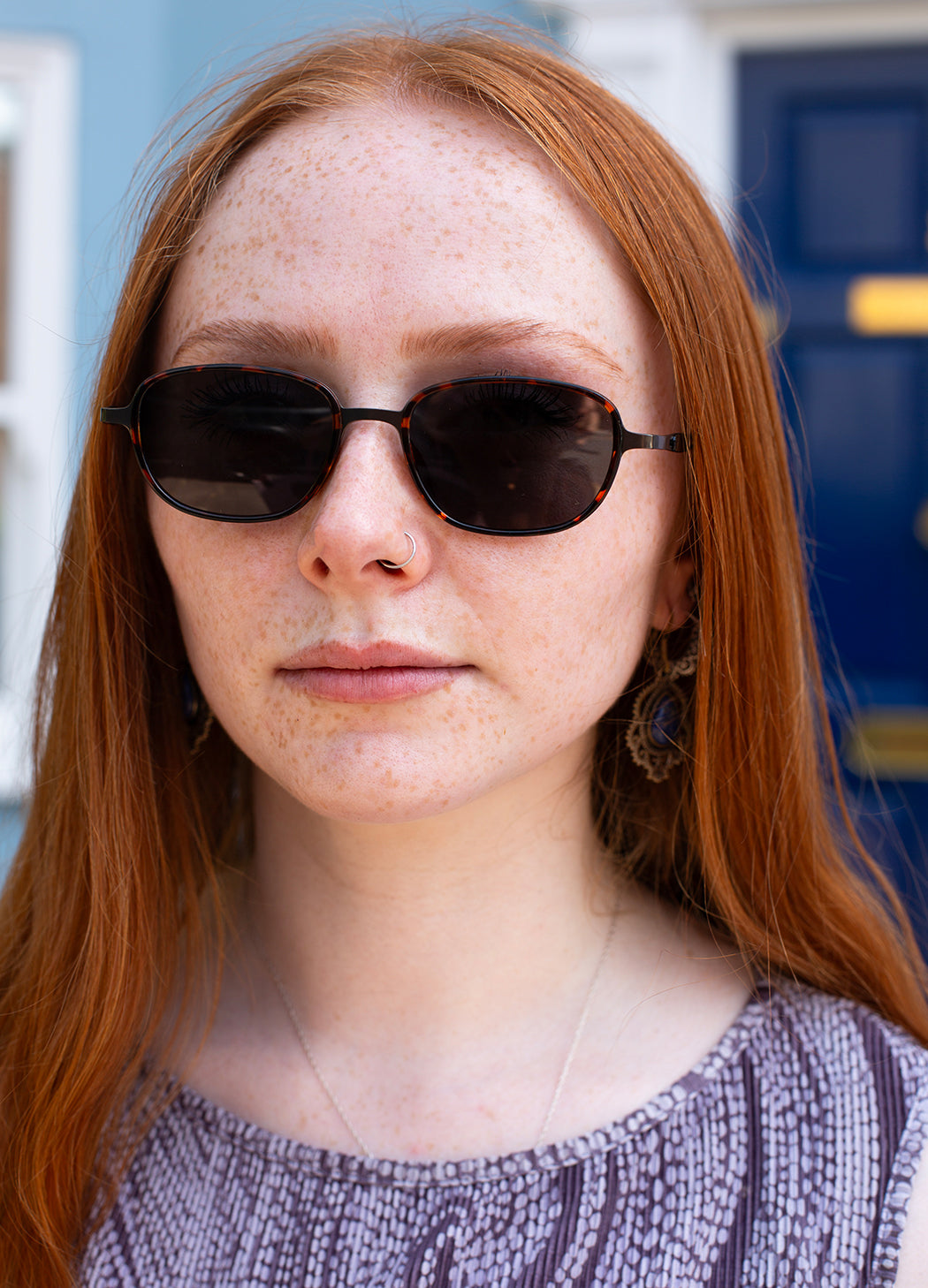 A close-up portrait of a woman with red hair and freckles wearing dark sunglasses and a textured purple top, set against a blurred blue door and wall background.