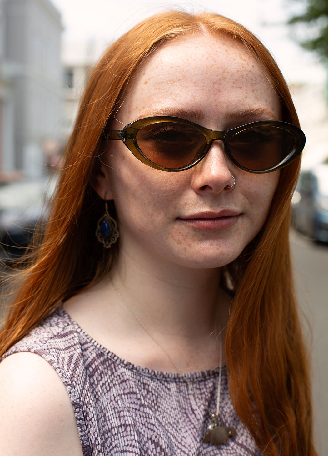 A close-up portrait of a woman with long red hair wearing dark brown cat-eye sunglasses, a textured lavender top, and dangling earrings, photographed outdoors on a blurred street background.