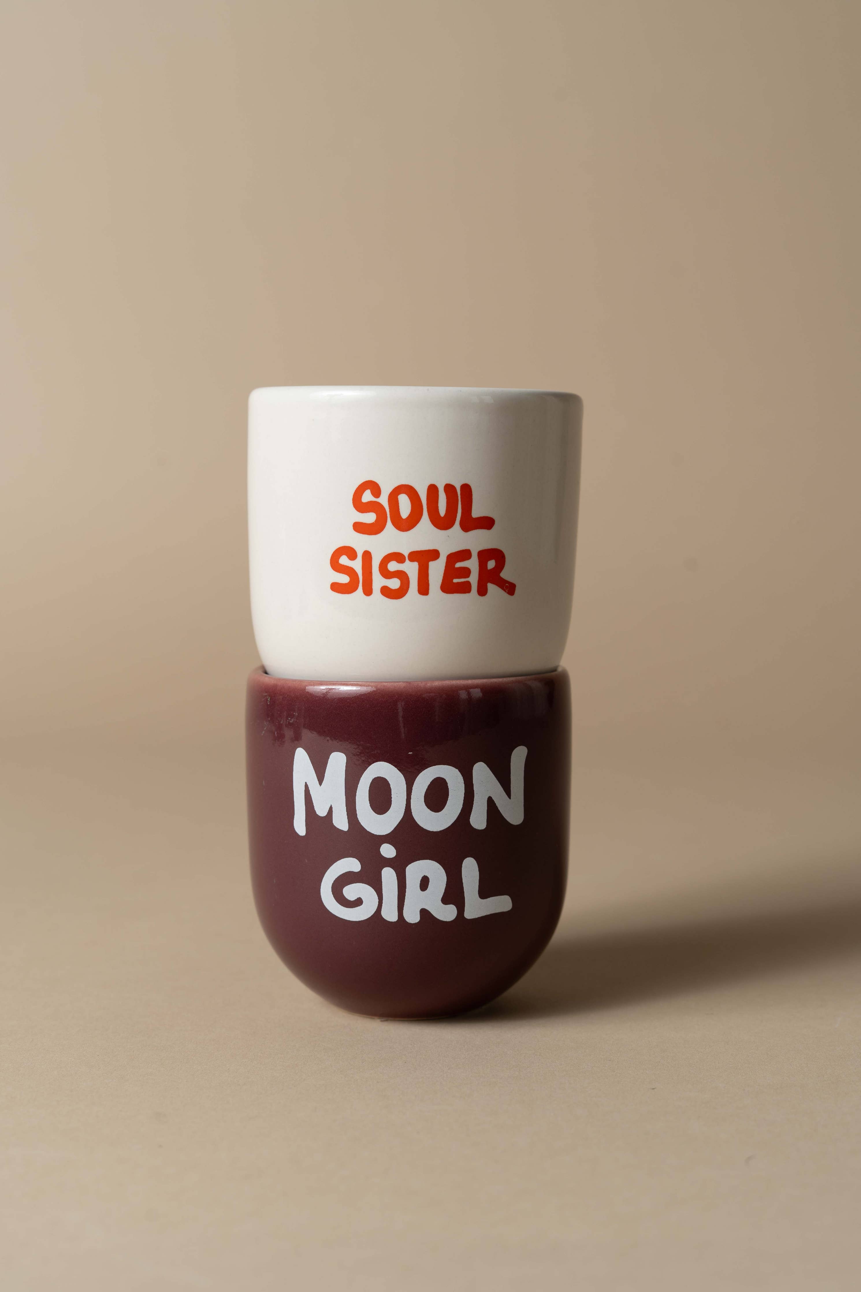 Two mugs stacked on a beige background, with 'Soul Sister' on the white mug and 'Moon Girl' on the brown mug.
