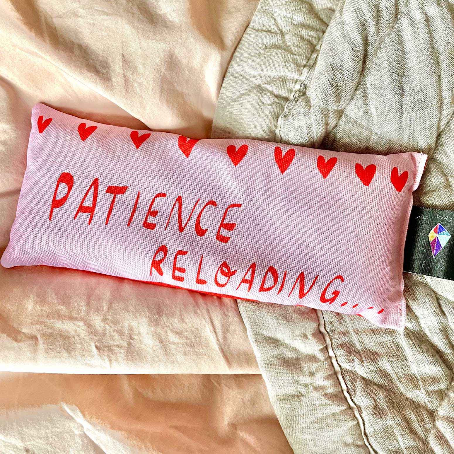 Pink eye pillow with 'PATIENCE RELOADING' text and red hearts on a beige fabric background