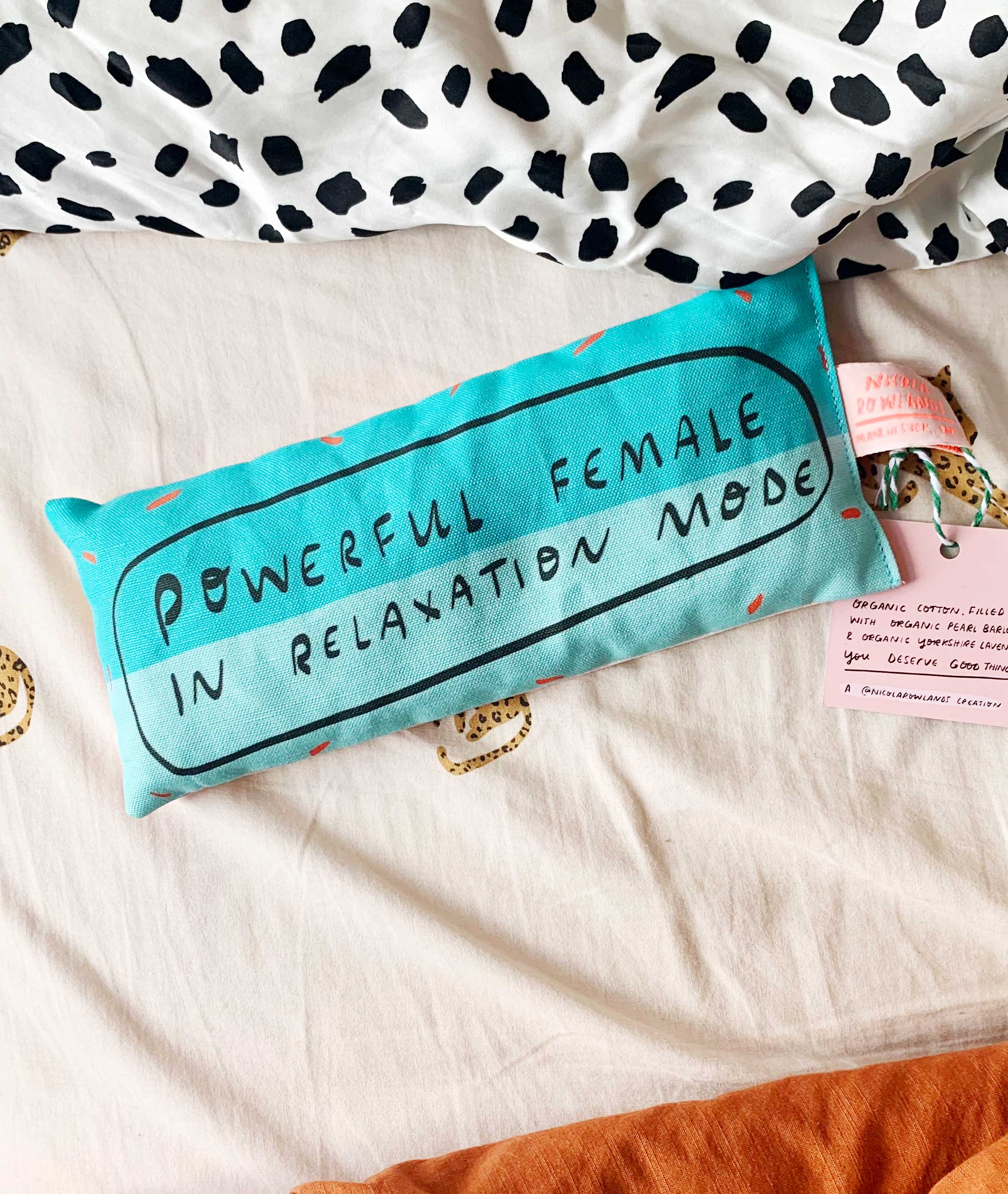 Turquoise pillow with 'Powerful Female in Relaxation Mode' text on a bed with polka dot blanket and accessories.