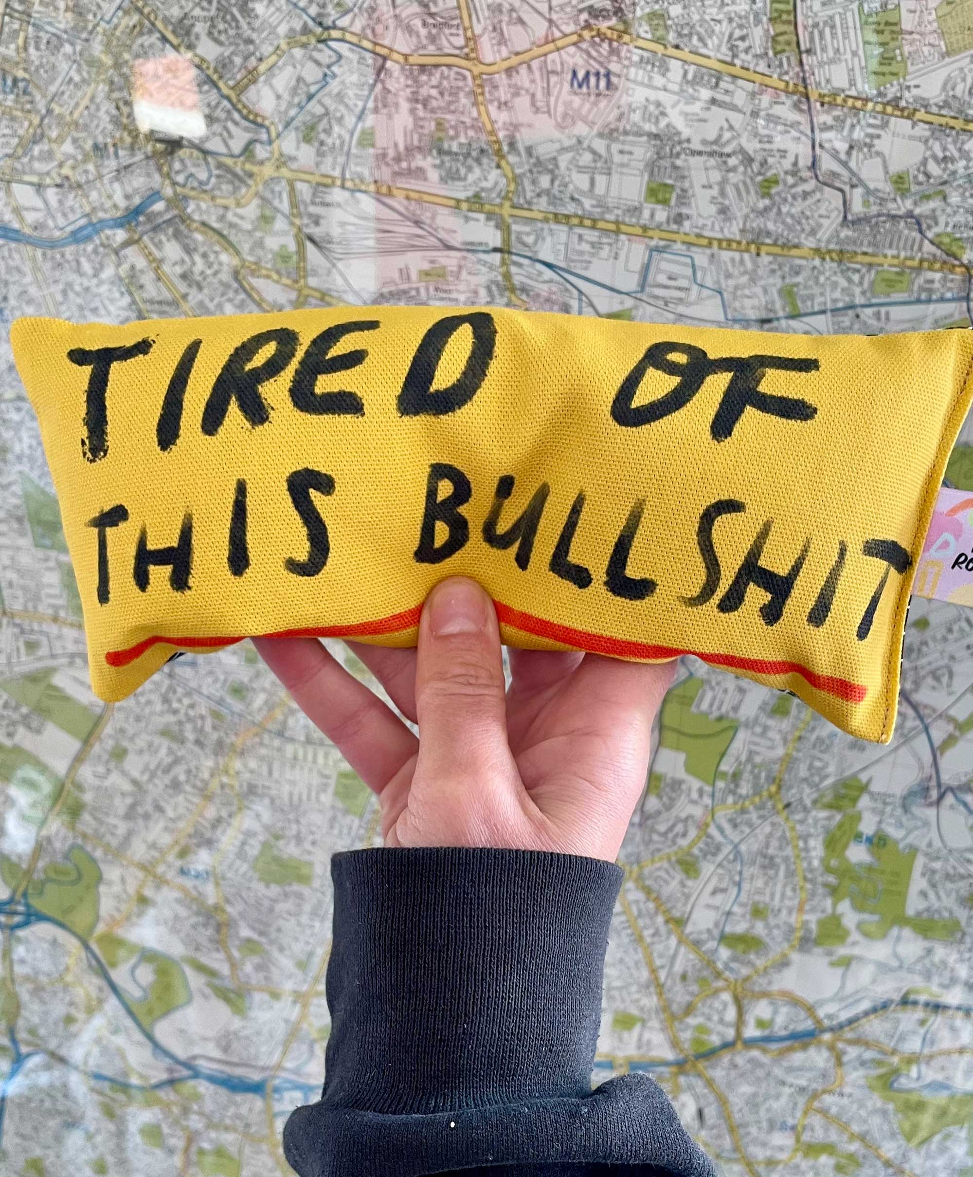 yellow eye bag pillow with the slogan tired of this bullshit on the front, held up against a map of Manchester where the item is hand made from organic materials